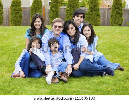 Large multiracial family of seven sitting together on lawn, dressed in blue colors. Five year old boy in front is disabled with cerebral palsy.