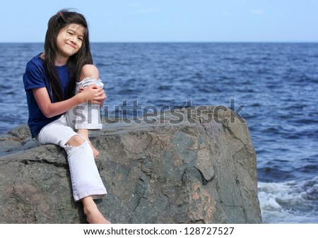 Little girl sitting on rock by lake shore, hugging bent knee