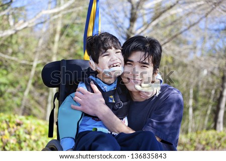 Little disabled boy in wheelchair hugging older brother outdoors, smiling together. Child has cerebral palsy.