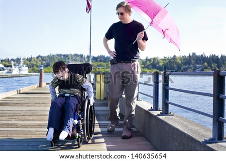 Father walking with disabled six year old son in wheelchair out on lake pier on sunny day