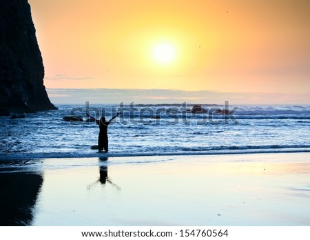 Silhouette of girl standing in waves, arms raised in praise to God at sunset