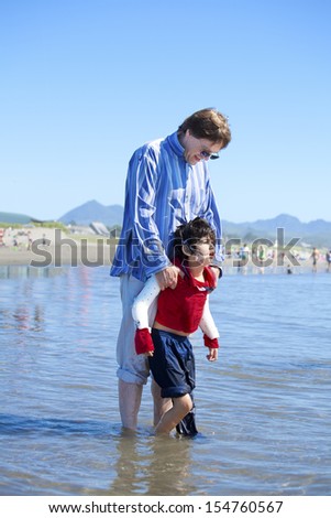Father helping disabled son walk in the ocean waves on beach. Child has cerebral palsy.