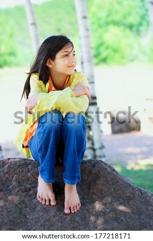 Young biracial girl quietly sitting on rock under trees outdoors in summer, peaceful scene