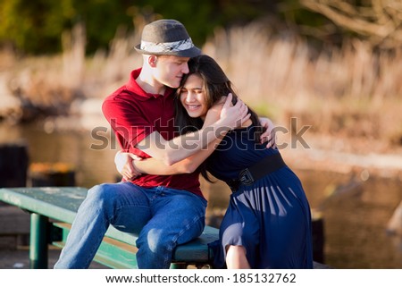 Young Caucasian man hugging his biracial girlfriend, sitting together on dock over lake