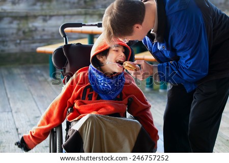 Father feeding disabled son a hamburger in wheelchair. Child has cerebral palsy