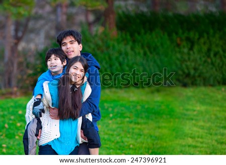 Disabled biracial child riding piggy back on his sister, family surrounding him, together at park.