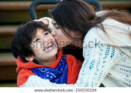 Big sister kissing disabled little brother seated in wheelchair on cheek