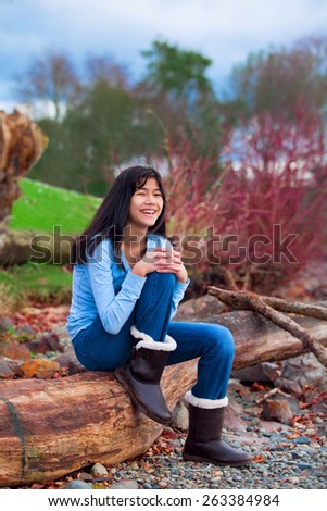Young biracial teen girl in blue shirt and jeans sitting on large log on rocky beach by lake