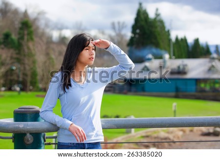 Young biracial teen girl standing, leaning against railing at park shading eyes to look off to side