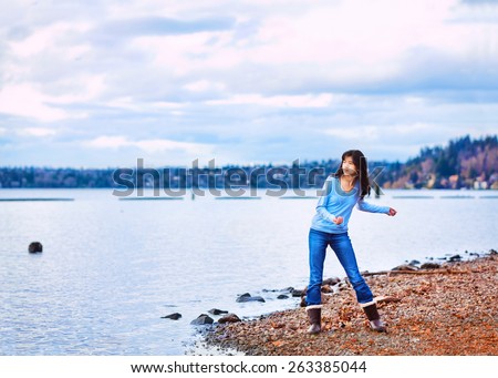 Young biracial teen girl in blue shirt and jeans along rocky lake shore, throwing rocks into the water on cloudy overcast day