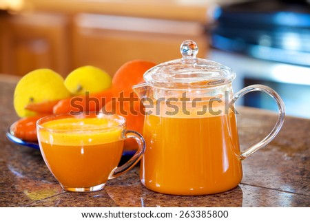 One cup of orange colored juice on kitchen counter with fruit and vegetables in background. Carrots, apples, lemons, oranges.