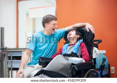 Father talking with disabled biracial son sitting in wheelchair while waiting in doctor\'s office, laughing together.