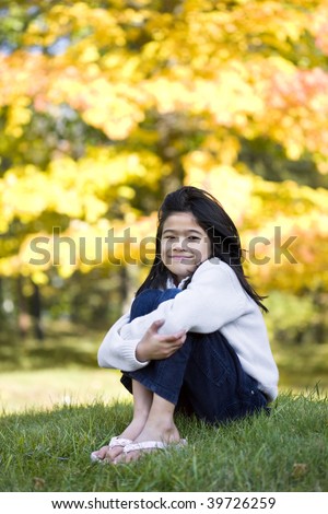 Little girl hugging knees sitting on lawn against bright autumn leaves in background