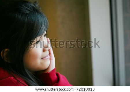 Little Asian girl quietly sitting by window, hand on chin