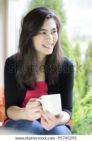 Beautiful biracial teenager or young woman drinking coffee next to window