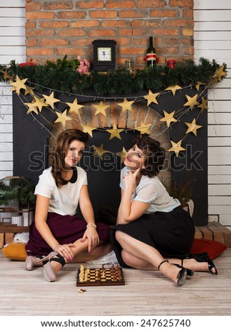 Two beautiful girl friends playing chess by the fireplace
