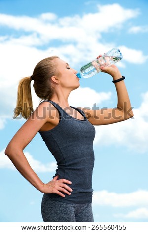 Fitness beautiful blonde woman drinking  water after sports on the background of sky and clouds