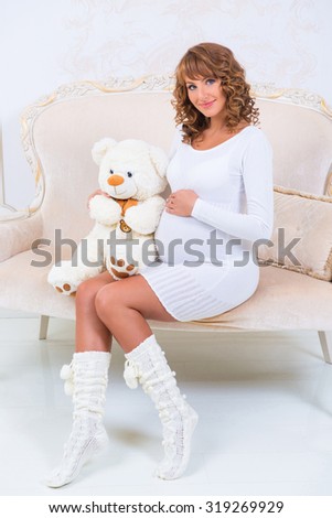 Beautiful pregnant girl in a knitted dress with teddy bear on the couch