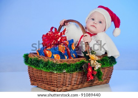 Baby Santa. Cute little girl dressed as Santa Claus sitting in a wicker basket with a gift. Basket decorated Christmas tree bells.