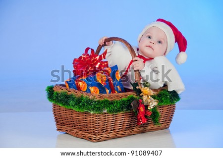 Baby Santa. Cute little girl dressed as Santa Claus sitting in a wicker basket with a gift. Basket decorated Christmas tree bells.