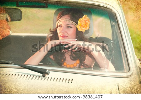 Beautiful brunette with flower in her hair sitting behind the wheel of an old car. Photos in the old style.