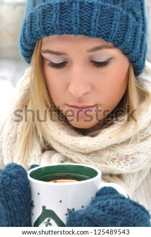 Photo of a beautiful blonde woman outside holding mug of hot tea in a winter clothing while snowing