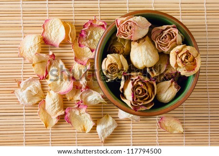 Dried rosebuds in clay tea cup and dry rose petals on a straw mat closeup top view