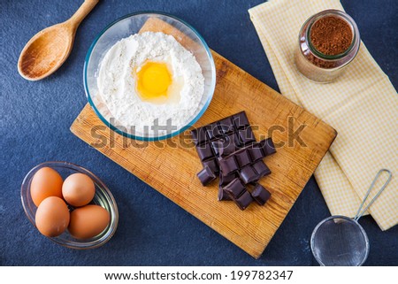 Baking ingredients for a chocolate cake - flour, eggs, chocolate, cocoa powder with a wooden spoon, sieve and napkin on a slate background