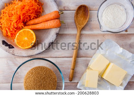Baking ingredients for a carrot cake - flour, butter, brown sugar, grated carrots, half an orange, a wooden spoon on a rustic wooden background