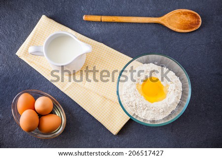 Baking ingredients - flour, milk and eggs with a wooden spoon and napkin on a slate background