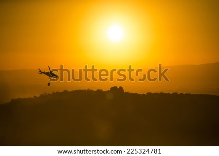 Fire fighting helicopter over burning forest at sunset.