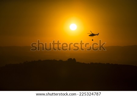 Fire fighting helicopter over burning forest at sunset.