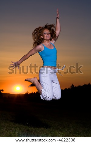 Friendly Redhaired woman in sportswear jumps up while being barefoot. Sun goes down in the background.