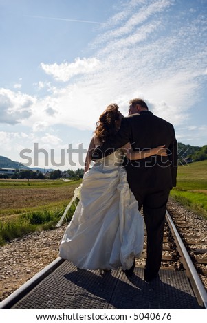 Bridal Couple (over 40 years old) amorous from the back. They are hugging each other while standing on a railway track. It is summer, great sky.
