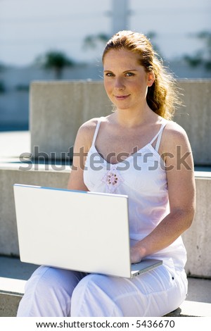 Working on her laptop. A woman (student?) sitting alone on a stair. Bright light and summery. Looking into camera.