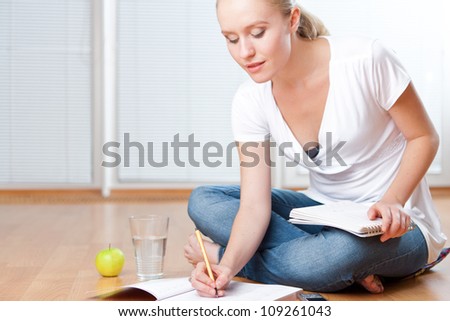 Closeup of young female student sitting on the floor and taking notes, studying and organizing with healthy snack including apple and fresh water