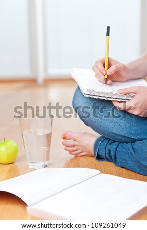 Closeup of young female student sitting on the floor and taking notes, studying and organizing with healthy snack including apple and fresh water