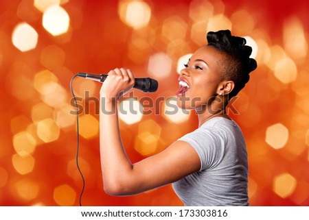 Beautiful teenager woman singing karaoke concert artist holding microphone, on red orange blurred lights background.
