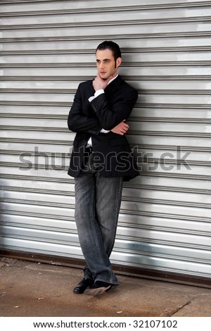 Handsome casual business man standing in front of and leaning against silver metal garage door with hand on chin thinking
