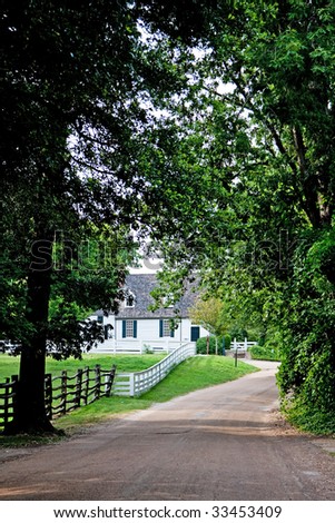 Driveway surrounded by trees next to meadow surrounded by a white fence leading towards a white colonial farm house