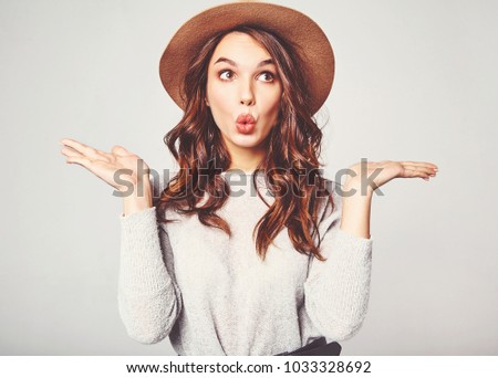 Comparison concept. Young brunette woman in casual clothes and brown hat displaying something on both flat hands for similar choice of product, gray background studio