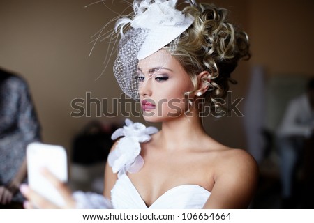 Happy beautiful bride blond girl in white wedding dress with hairstyle and bright makeup on home background looking in the mirror