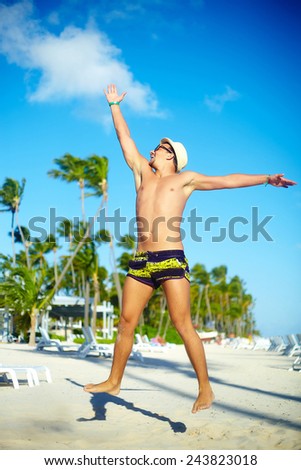 Happy handsome muscled man in sunhat on beach jumping behind blue sky behind blue sky