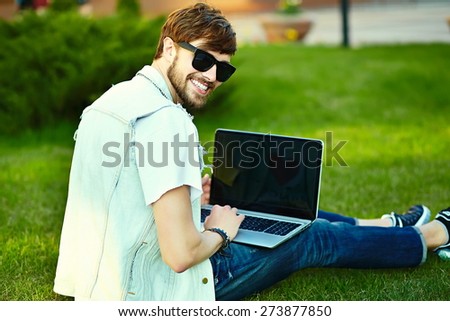 Funny smiling hipster handsome man guy in stylish summer cloth in the street sitting on grass in the park with notebook