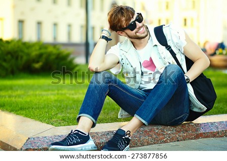 Funny smiling hipster handsome man guy in stylish summer cloth in the street sitting on grass in the park