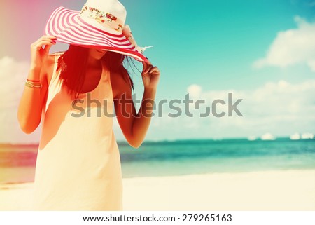 Hot beautiful woman in colorful sunhat and dress walking near beach ocean on hot summer day on white sand