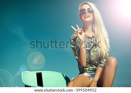 High fashion look.glamor stylish sexy beautiful young blond model girl in summer bright casual hipster clothes with skateboard behind blue sun sky showing her tongue and peace sign