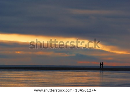 Two people walking on a beach towards the sun at sunset. Orange and blue colored clouds and an opening in the clouds formed a spectacular view of the sunset.