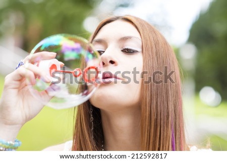 Closeup portrait of beautiful young woman inflating colorful soap bubble, outdoor.