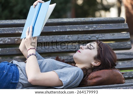 Beautiful girl lying on old wood bench reading blue book in the park, education lifestyle concept.